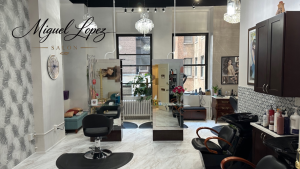 Elegant hair salon interior with black styling chairs, large mirrors, chandeliers, and a windowed reception area.